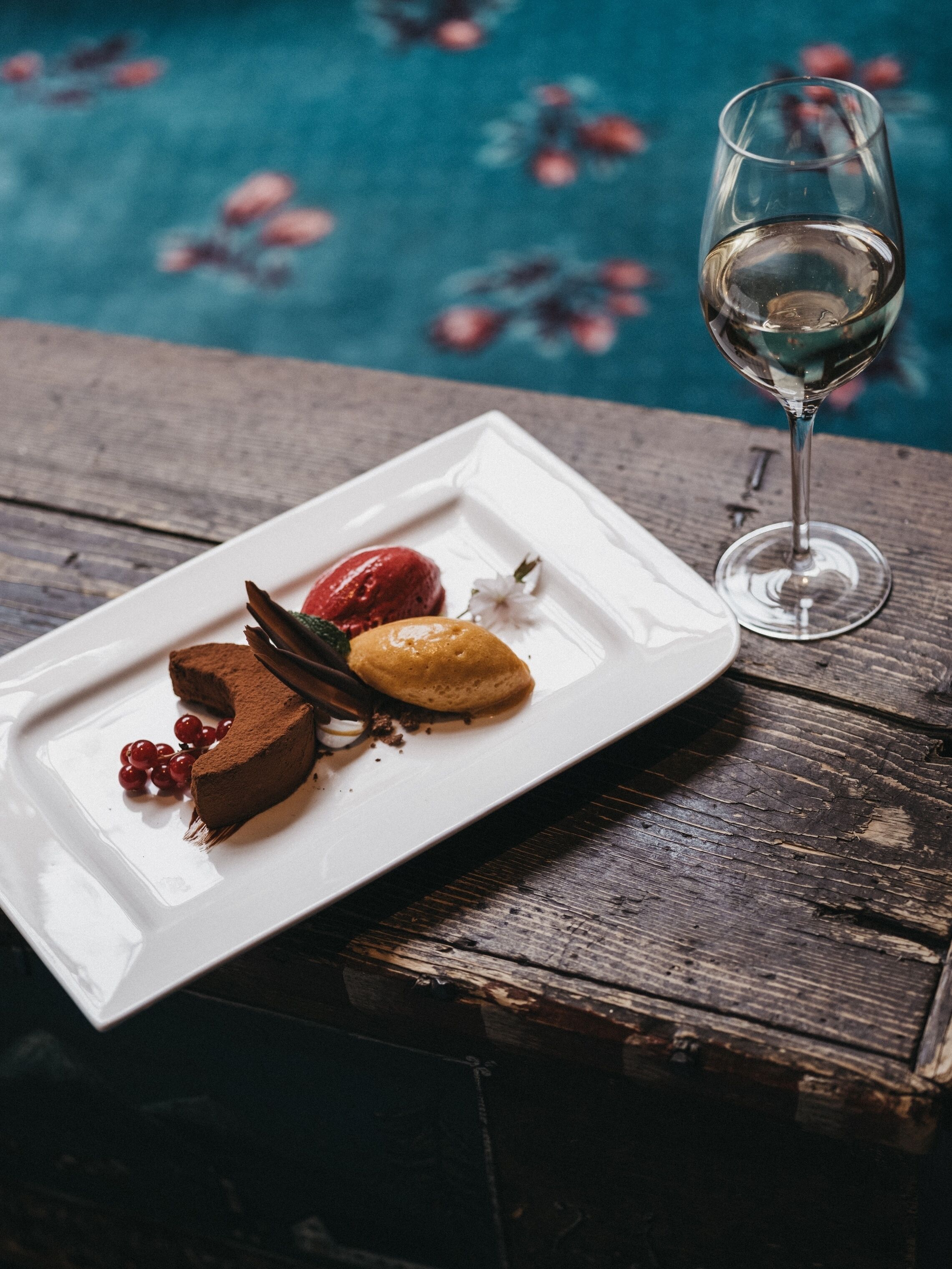 A dessert plate with chocolate cake, berries, and a scoop of ice cream on a white plate, accompanied by a wine glass, set on a wooden table with a pool in the background.