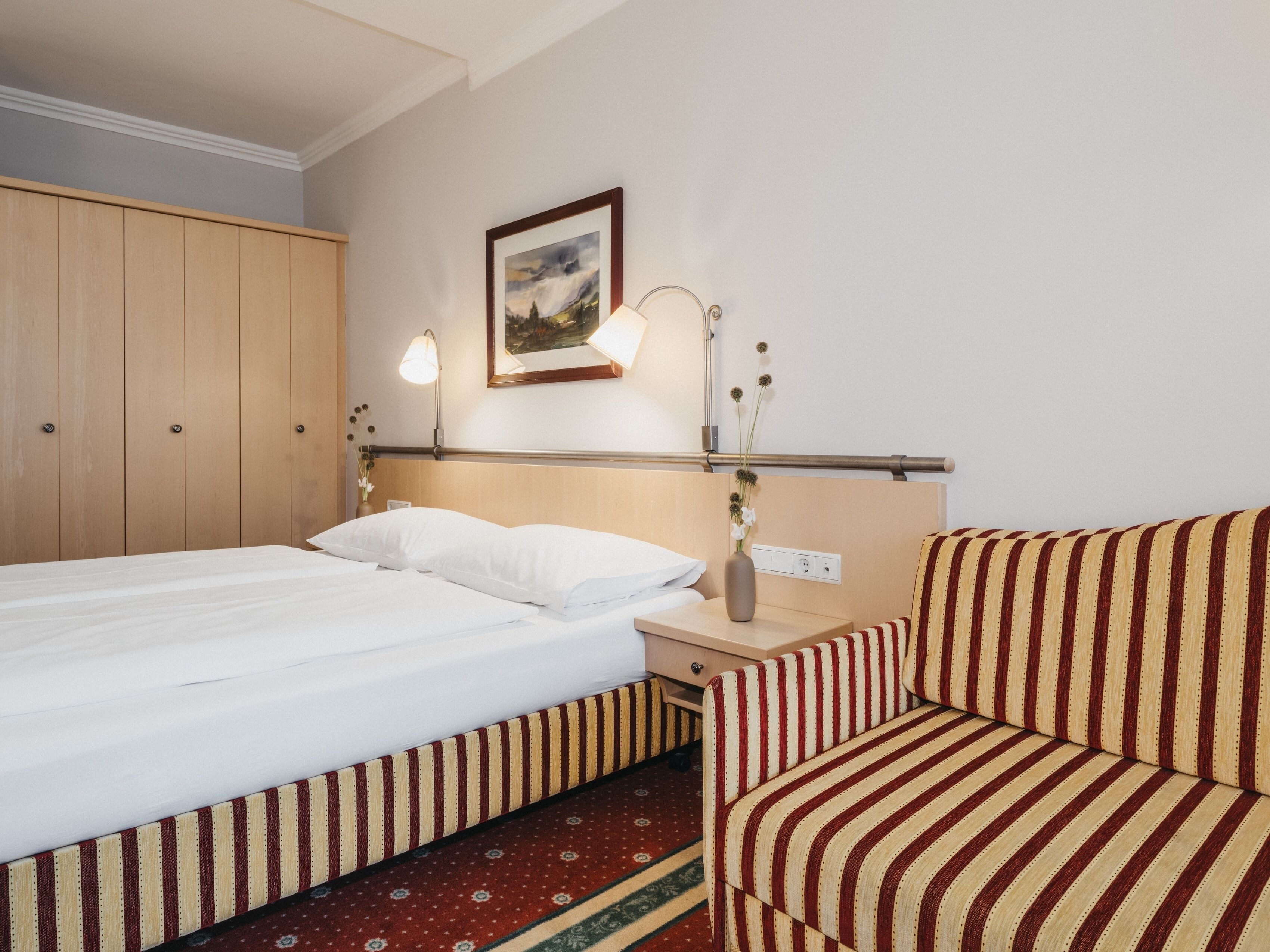 A hotel room with a bed, a bedside table with a lamp, a chair, and a painting on the wall.