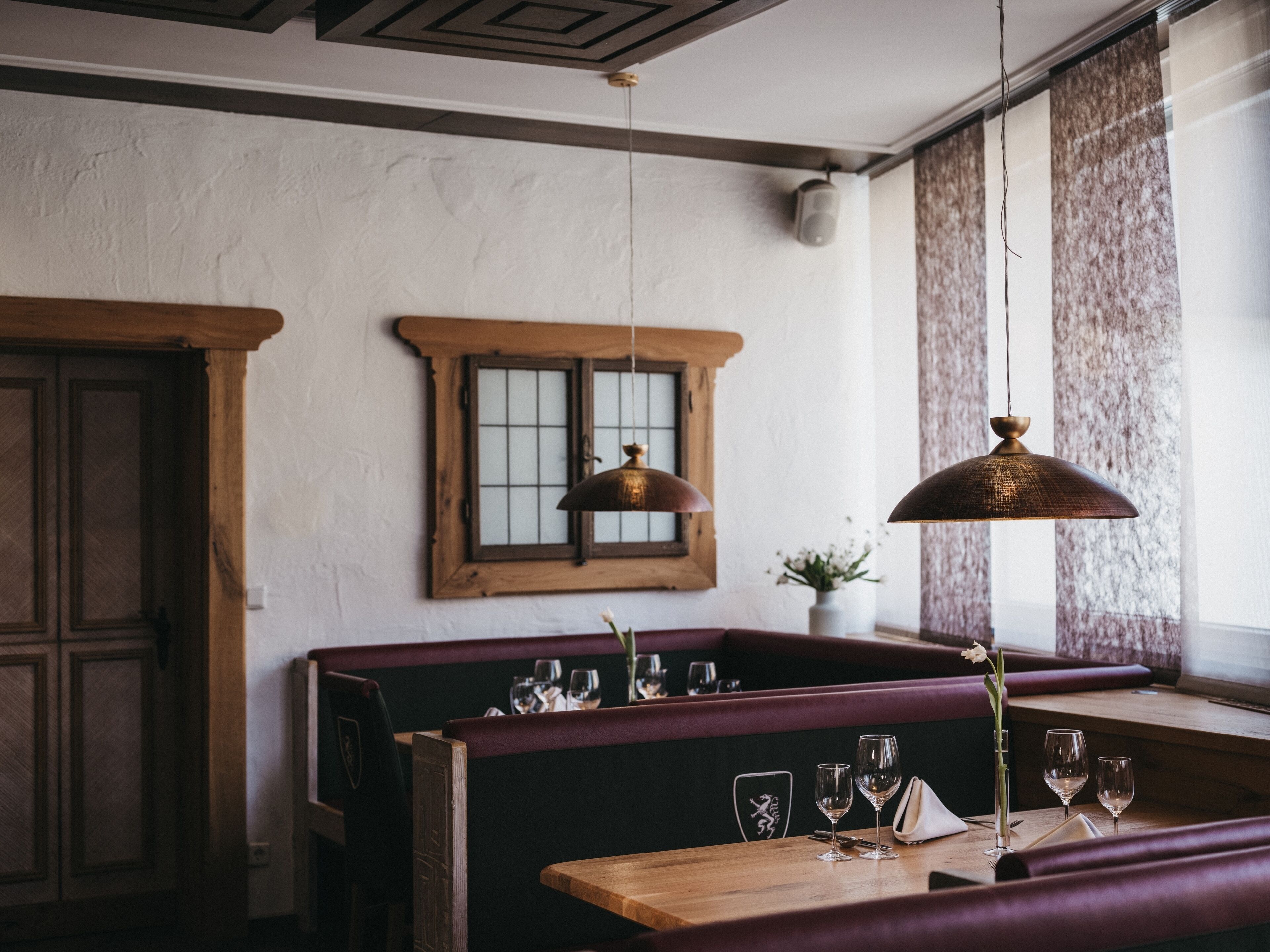 Interior de un restaurante con bancos de madera, una mesa con copas de vino y una lámpara colgante.