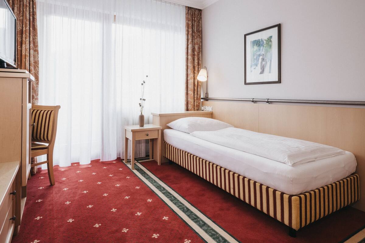 A hotel room with a single bed, a wooden nightstand, a chair, a desk, and a framed picture on the wall.