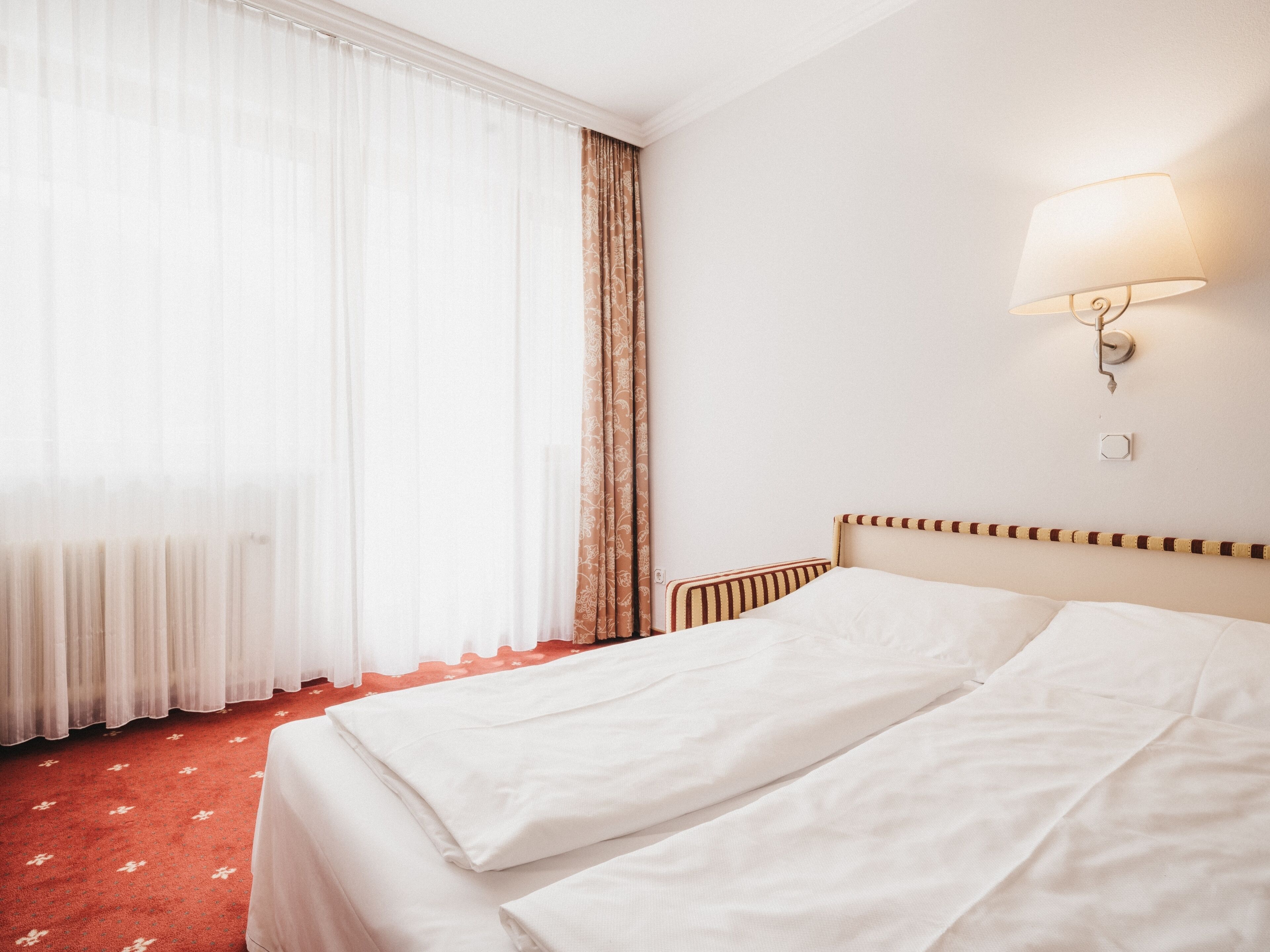 Habitación de hotel con cama, ventana con cortinas translúcidas, lámpara fijada a la pared y radiador.