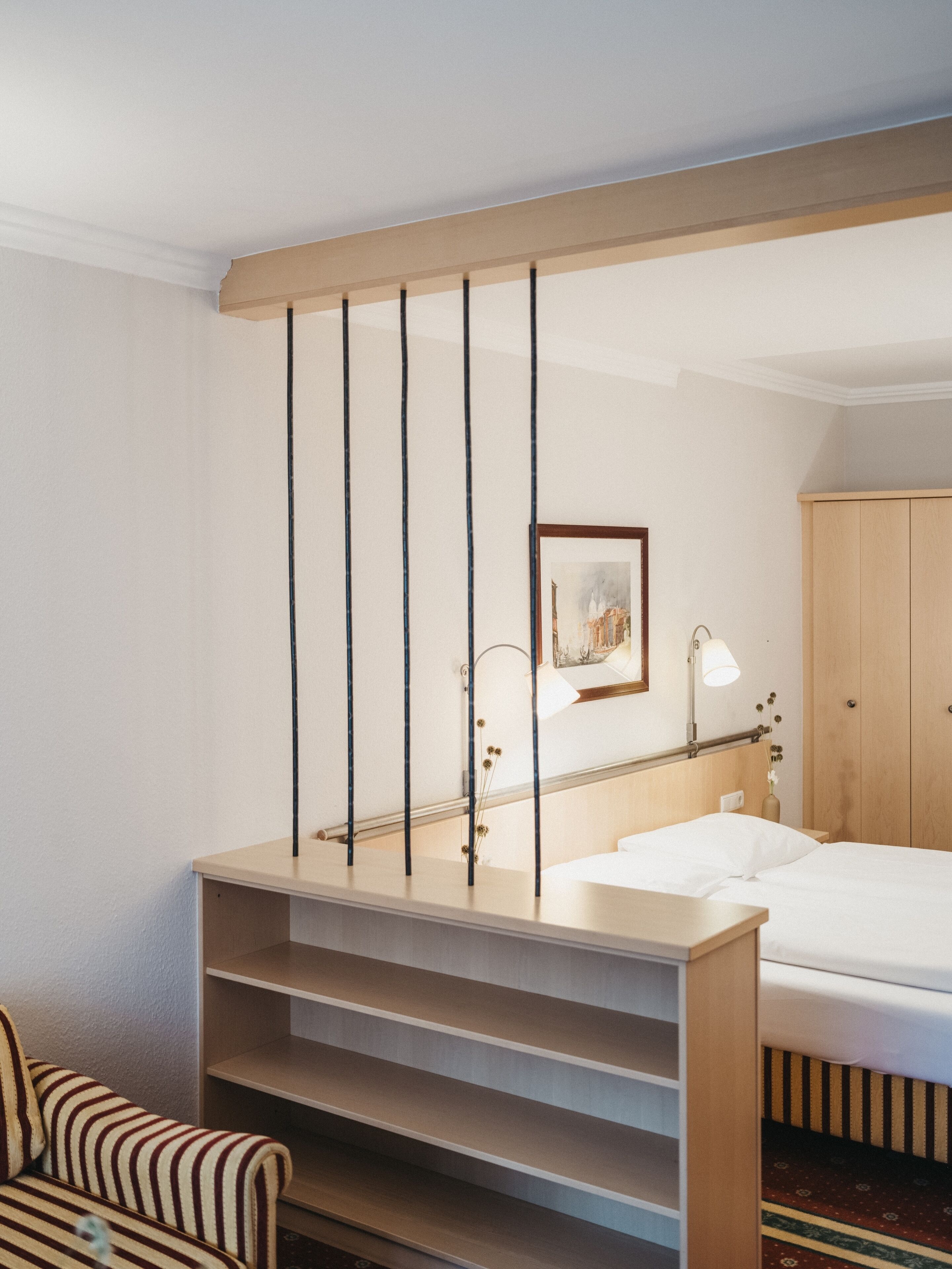 A hotel room with a bed, a desk, a chair, a wardrobe, and a painting on the wall.