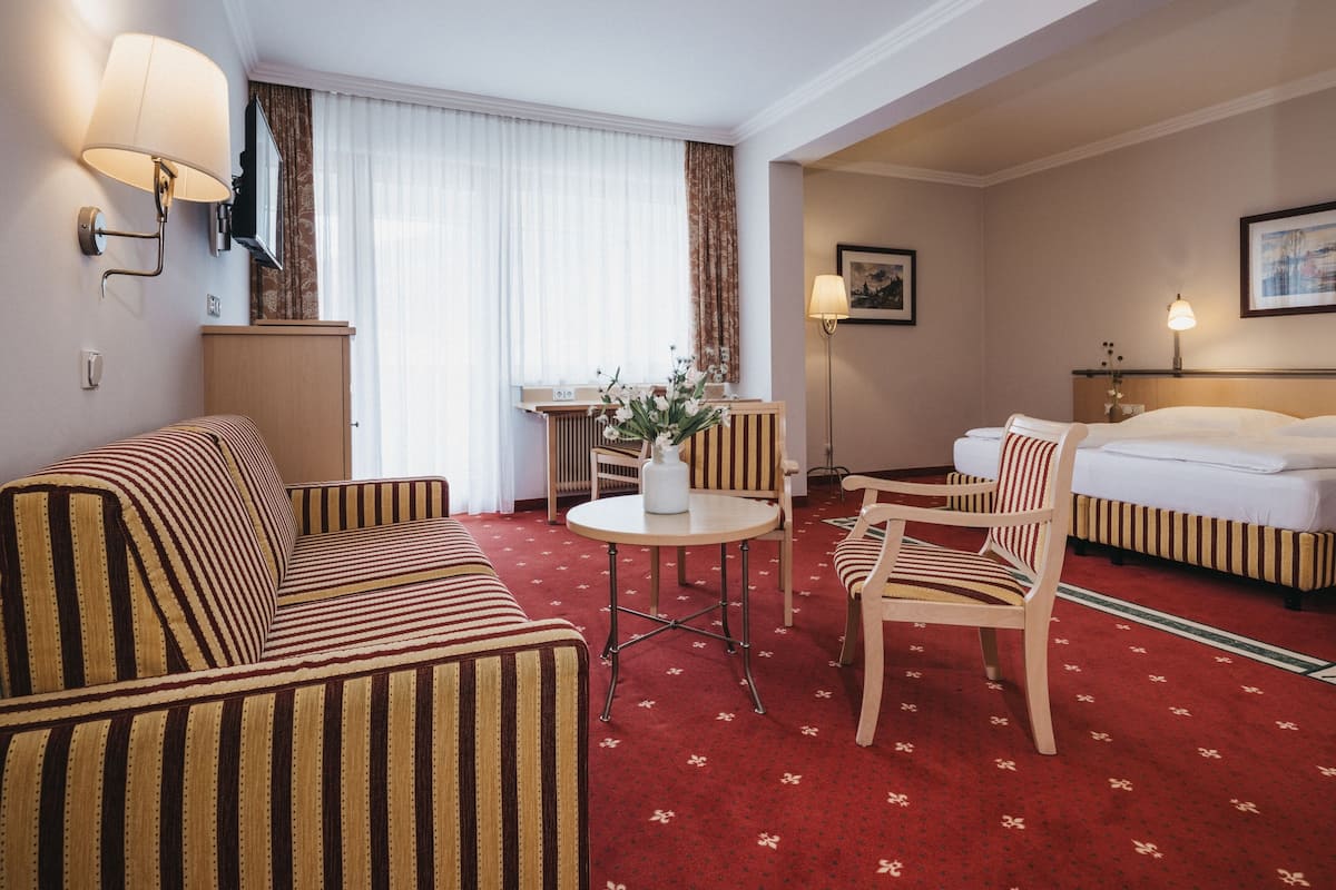 A hotel room with a striped sofa, a round table with a vase of flowers, a wooden chair, a bed, and a window with curtains.
