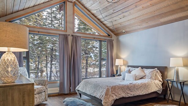 Ein Schlafzimmer mit einem großen Bett, zwei Nachttischen, einem Sessel und Blick auf Bäume durch große Fenster.