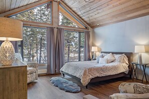 A bedroom with a large bed, two bedside tables, a chair, and a view of trees through large windows. - Lovely Suite with Full Kitchen, Close to Downtown! (Canmore)