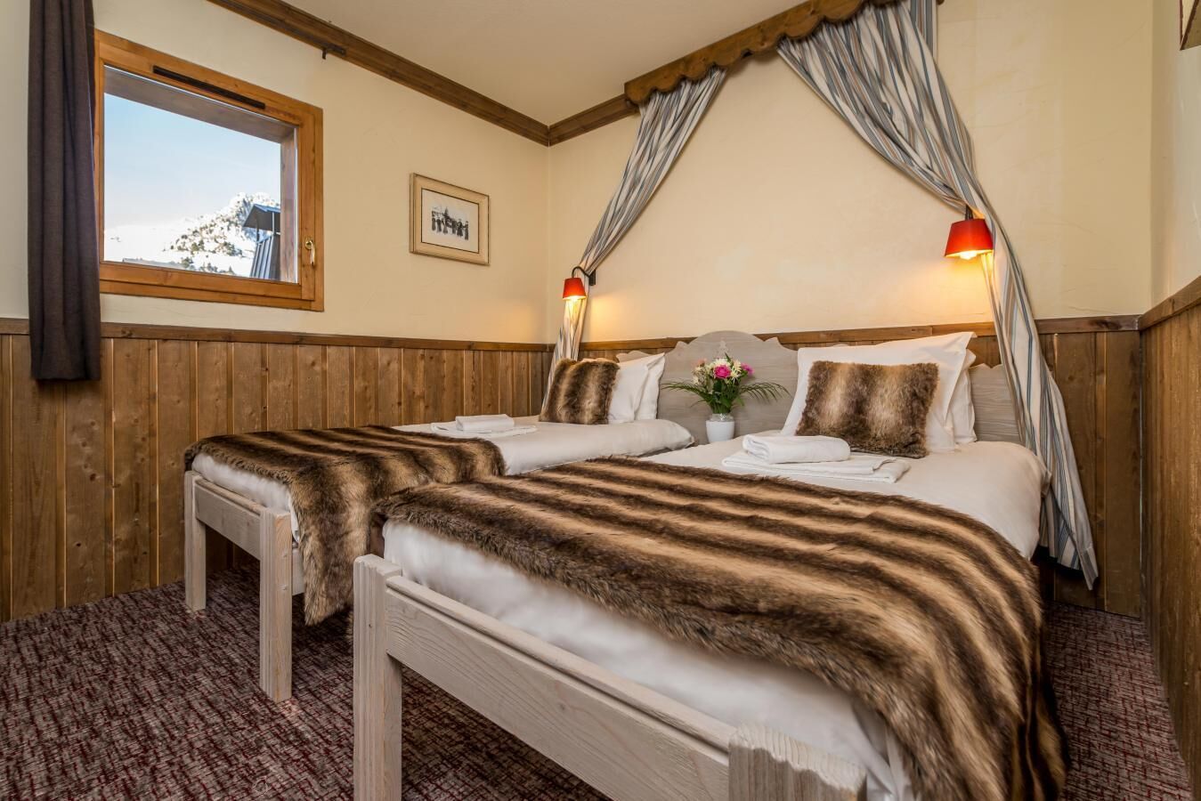 A double bed with a fur throw, a bedside table, a window with curtains, and a wall-mounted lamp.