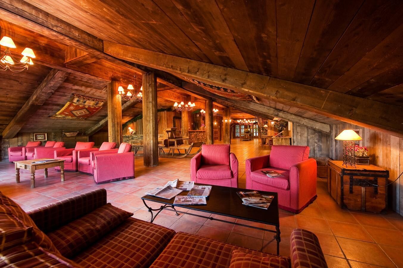 An attic-style room with wooden beams, red upholstered sofas, a coffee table, and a chandelier.