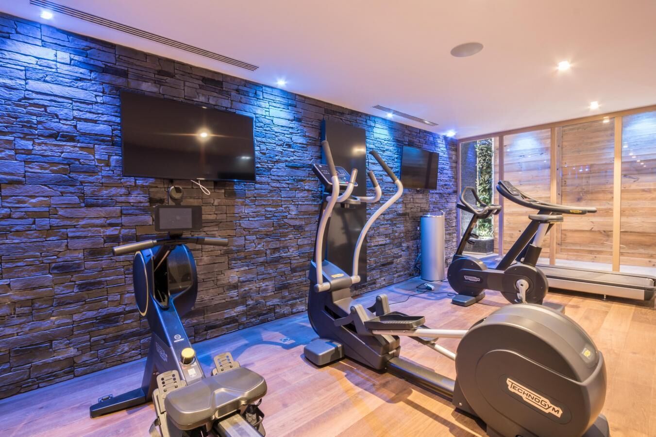 Ein Heimfitnessraum mit Steinwand, Fernseher und Trainingsgeräten, darunter Laufband, Crosstrainer und Indoor-Fahrrad.