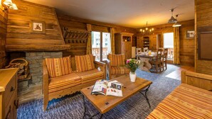 Une pièce chaleureuse, aménagée dans le style d’un chalet en rondins, avec une cheminée en pierre, un canapé à rayures, une table à manger avec des chaises et une vue sur l’extérieur grâce à de grandes fenêtres.