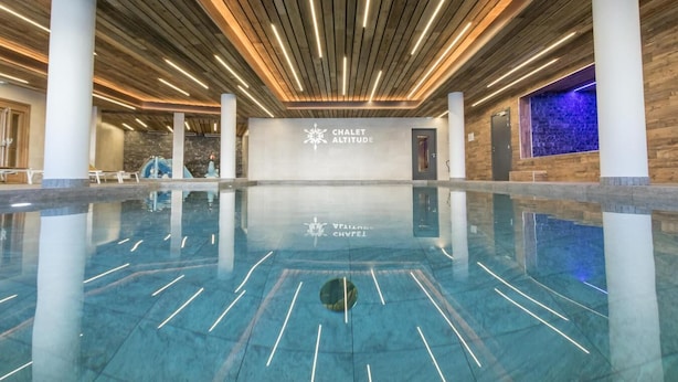 Une piscine intérieure spacieuse au design moderne, avec un plafond en bois et une enseigne indiquant « CHALET ALTITUDE ».