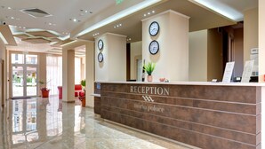 En receptionsdisk med ure, et skilt der siger "RECEPTION maria palace", og en potteplante.