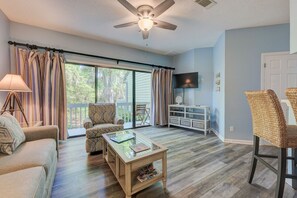 Un salon avec un ventilateur de plafond, un canapé, un fauteuil à motifs, une table basse en verre et un téléviseur fixé au mur.