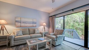 Ein Wohnzimmer mit Sofa, Sessel, Couchtisch und Lampen. Ein Gemälde hängt an der Wand, und durch ein großes Fenster ist eine Baumlandschaft zu sehen.