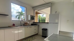 Cozinha com armários brancos, micro-ondas, geladeira, janela com vista para palmeiras e uma pequena lixeira.