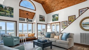 A spacious living room with a high ceiling, large windows overlooking a body of water, and a cozy seating area with a sofa and armchairs.
