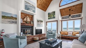 A spacious living room with a fireplace, comfortable seating, large windows, and artwork on the walls.