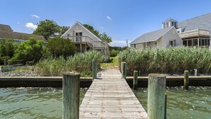 A wooden dock leading to a house with a balcony, surrounded by water and vegetation.