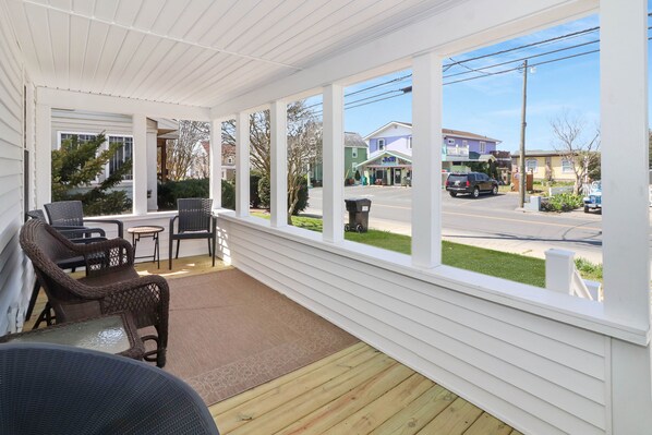 A covered porch with wicker furniture, a glass table, and a view of a street with houses and a store. - Stella del Mare | Remodeled South Main Cottage Walk Downtown w/ Patio Dining, King Suite & Charm (Chincoteague Island)