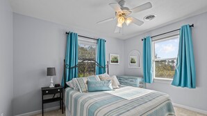 Ein Schlafzimmer mit einem Bett, einem Nachttisch, einem Deckenventilator und zwei mit Vorhängen versehenen Fenstern.
