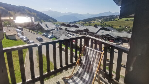 Un balcon avec une chaise longue à rayures, une vue sur un village aux bâtiments traditionnels et, au loin, une chaîne de montagnes.