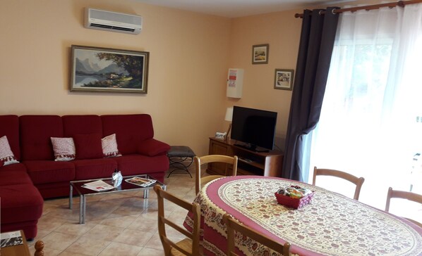 A living room with a red sofa, a coffee table, a TV, a dining table with a patterned tablecloth, and a painting on the wall.