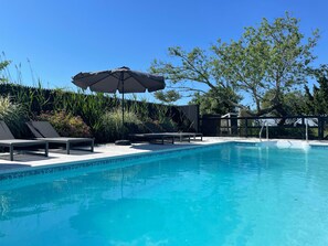 En klarblå pool med solstolar och ett parasoll, omgiven av grönska och ett staket.