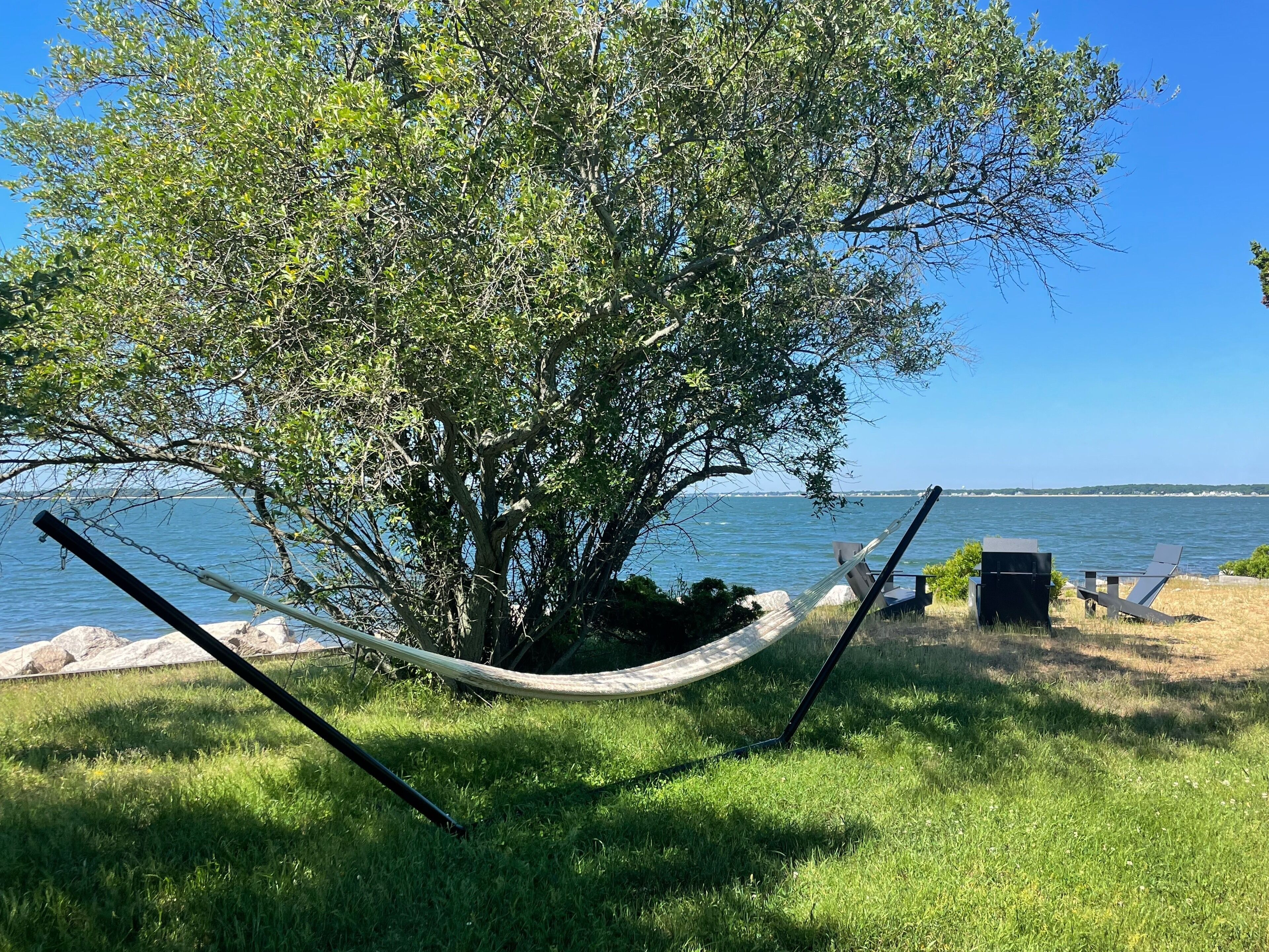 Una hamaca atada entre dos árboles a orillas de un lago, con una silla y una parrilla cerca.