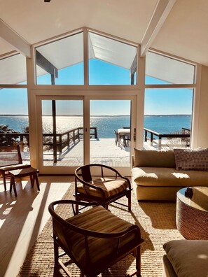Ein geräumiges Zimmer mit großen Fenstern und Blick auf einen Strand, ausgestattet mit einer Couch, Sesseln und einem Essbereich.