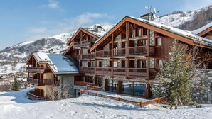 A multi-story wooden chalet with balconies, set against a snowy mountain backdrop.