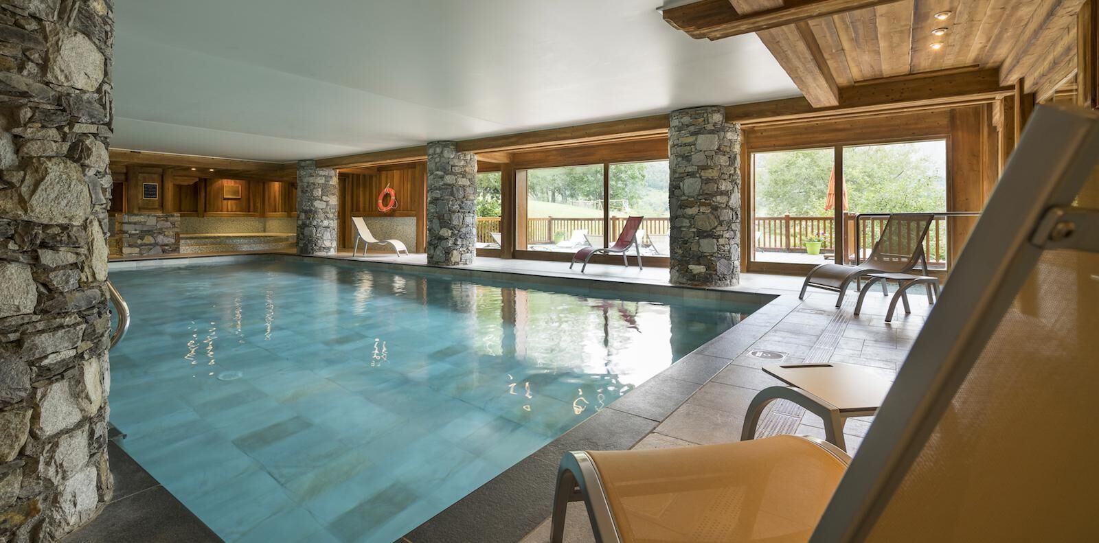 A large indoor swimming pool with stone pillars, wooden deck chairs, and large windows offering views of the outdoors.