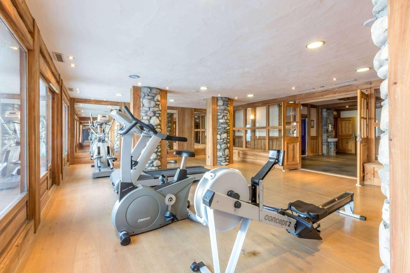 Een fitnessruimte met cardioapparatuur, houten vloeren, stenen zuilen en grote ramen.