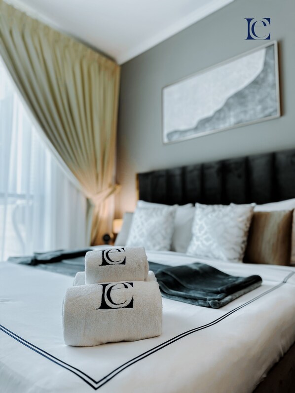 A neatly made bed with a grey headboard, white pillows, and a black throw blanket. There are two towels with a logo on them placed on the bed. A painting is hanging on the wall above the bed.