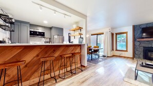 A modern kitchen with a wooden bar, grey cabinets, and a stone fireplace. - Pet-Friendly, Bright Modern Gem, Gas Grill, Near Trails, Quiet Location (Mammoth Lakes)