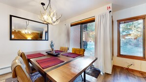 A dining area with a wooden table, chairs, a chandelier, and large windows with curtains. - Pet-Friendly, Bright Modern Gem, Gas Grill, Near Trails, Quiet Location (Mammoth Lakes)