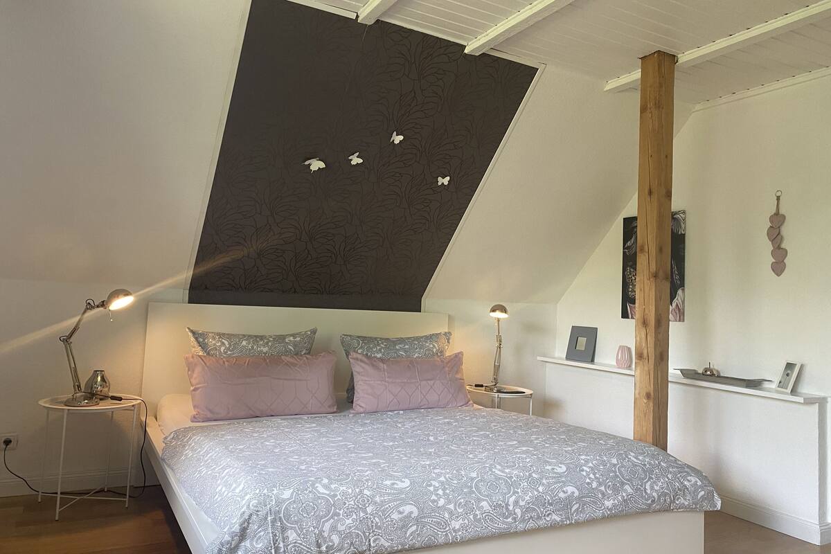 Ein Schlafzimmer mit schrägverdeckter Decke, einem Bett mit gemusterten Textilien, zwei Nachttischlampen und einem kleinen Tisch mit Lampe.