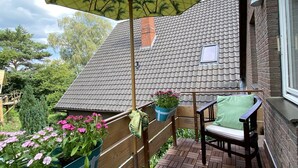 A wooden deck with a tiled floor, a table with a cushion, a chair, a hanging umbrella, and potted plants. - Worpsweder Blick (Osterholz-Scharmbeck)