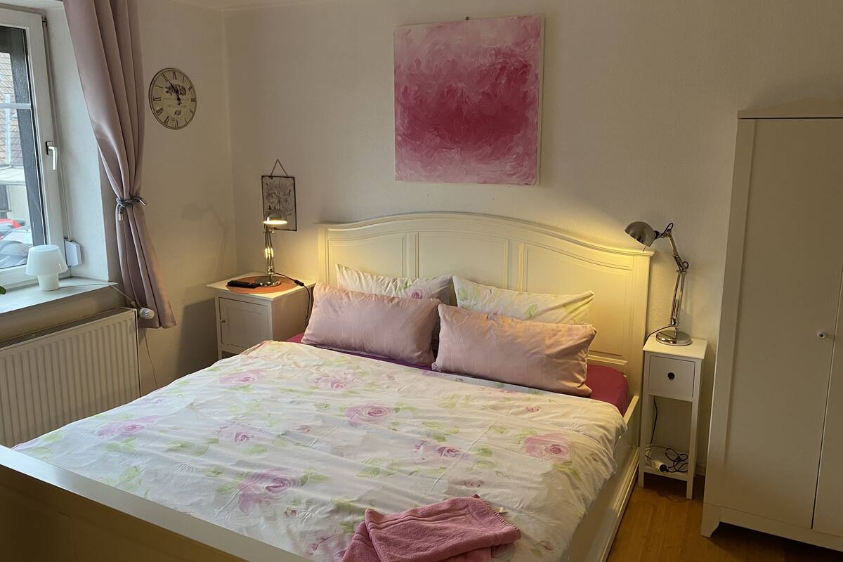 Ein Schlafzimmer mit Bett, Nachttisch, Lampe, Heizkörper, Fenster mit Vorhängen und einer Wanduhr.