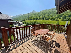 Eine Holzterrasse mit Tisch und Stühlen, mit Blick auf eine grüne Landschaft und Berge im Hintergrund.