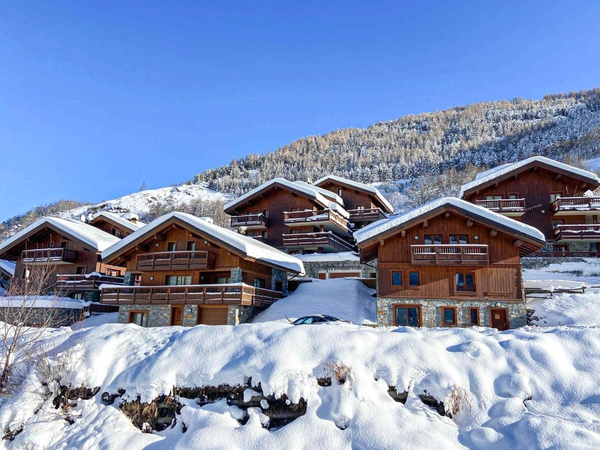 A cluster of wooden chalets with balconies, set against a snowy mountain backdrop.