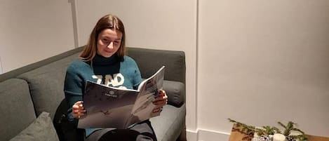 Una persona sentada en un sofá gris, leyendo un libro. Hay una mesa de centro de madera con una decoración de rama de pino.