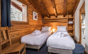 Une chambre de style cabane avec deux lits, des murs en bois, une fenêtre avec des rideaux, une affiche au mur et une étagère avec des serviettes.