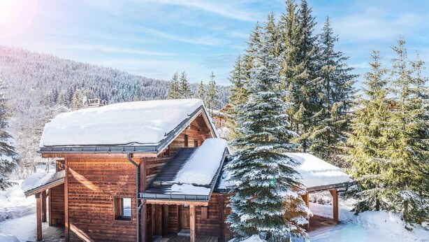 Une cabane en bois, dont le toit est recouvert de neige, entourée d’arbres à feuilles persistantes dans un paysage enneigé.