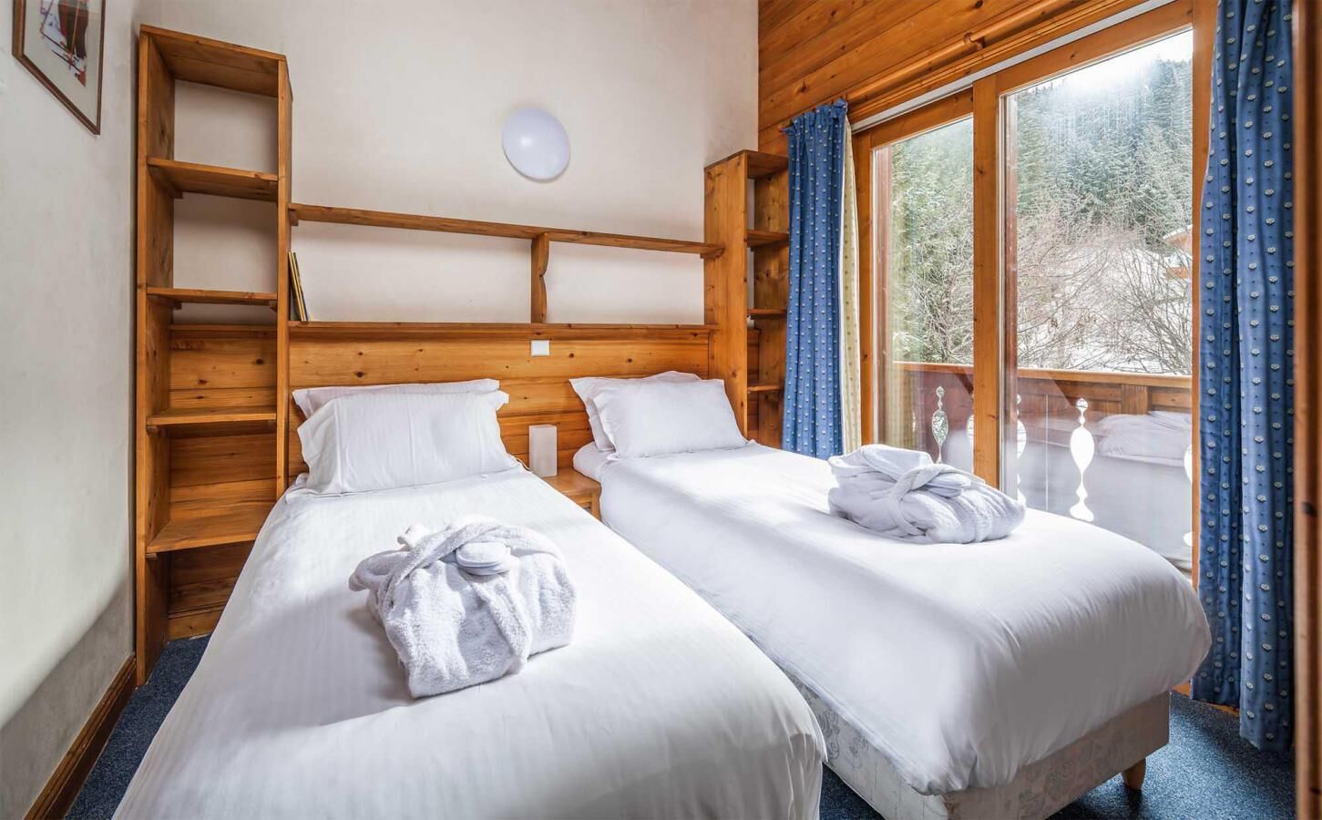 A room with two beds, wooden headboard, a shelf unit, and a large window with curtains.