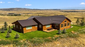 A two-story wooden cabin with a patio, surrounded by a grassy landscape and distant hills.