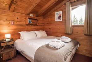 A wooden cabin bedroom with a bed, a nightstand, a lamp, and a window with curtains. - Chalet, terrace, fireplace or stove, parking, tv, ski locker, 165m², La Tania (Courchevel)