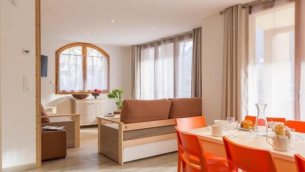 A modern dining area with a wooden table and orange chairs, a sofa, and a kitchen area with a window and curtains. - 50m from ski slopes, balcony, tv, ski locker, 84m², Serre Chevalier (Briançon)