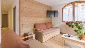 A modern living room with a wooden wall panel, a sofa, a TV mounted above it, a window with curtains, and a table with a plant. - 50m from ski slopes, balcony, tv, ski locker, 84m², Serre Chevalier (Briançon)