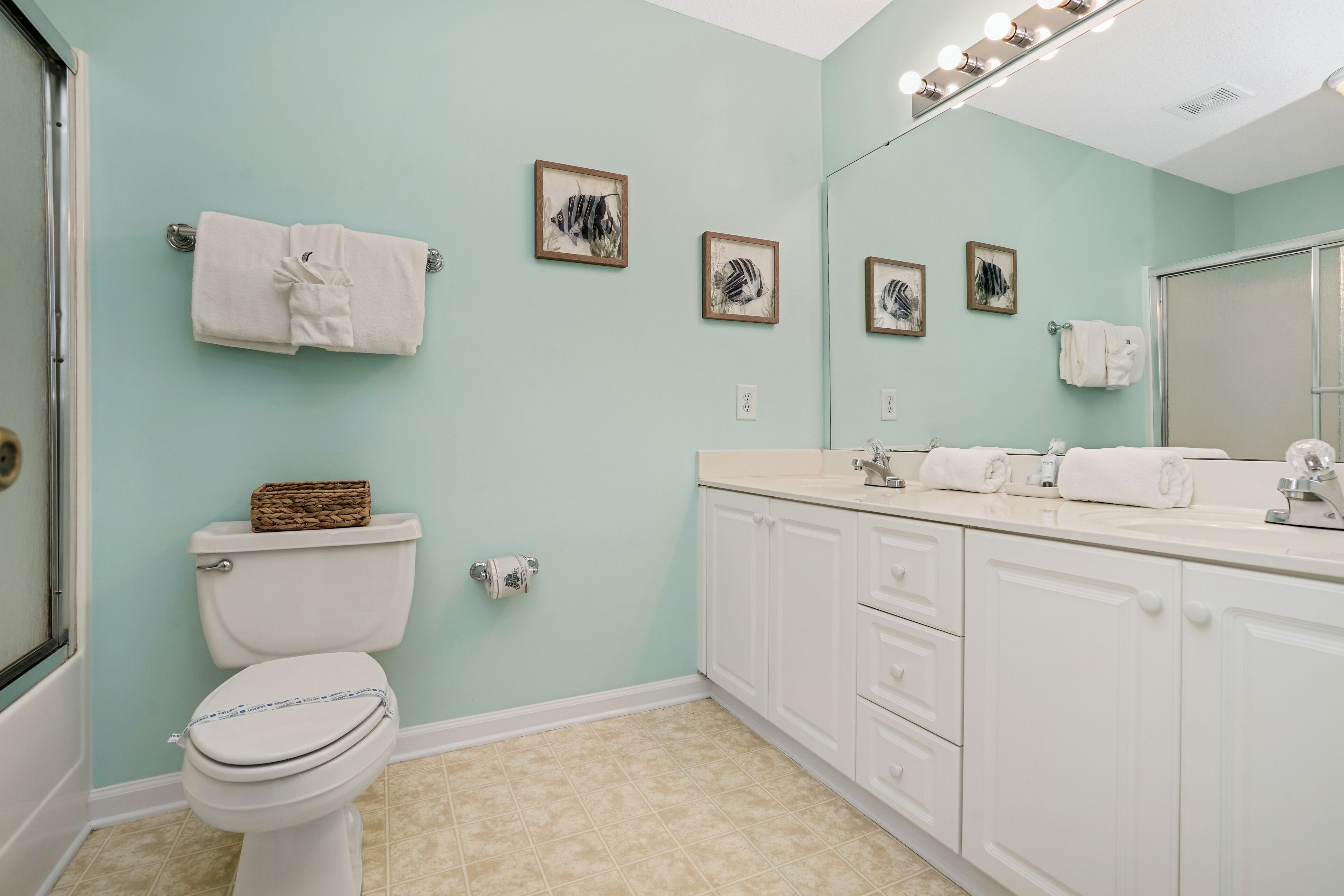 Une salle de bain avec des toilettes, un lavabo et un miroir. Des serviettes sont accrochées au mur et un panier est posé sur les toilettes.