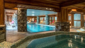 An indoor swimming pool with stone pillars, lounge chairs, and a view of the outside.