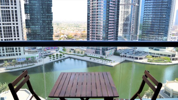 Vista desde un balcón con una mesa y sillas de madera, con vistas a una masa de agua y un paisaje urbano con edificios altos.